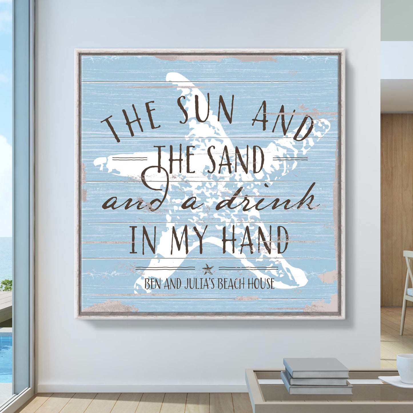 The image shows a coastal-themed wall art piece with a distressed blue wood background and a white starfish design. The text reads: "The sun and the sand and a drink in my hand" followed by "Ben and Julia’s Beach House." The artwork has a relaxed, beachy vibe, perfect for coastal or vacation home decor.