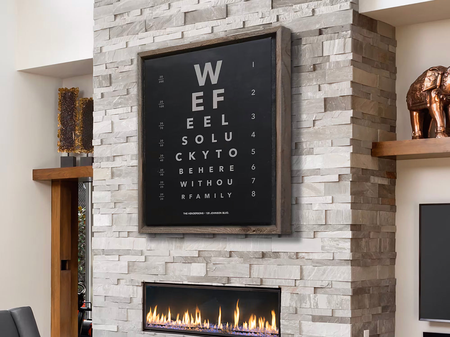 This image shows a stylish living room with a modern stone fireplace and a unique wall art piece above it, designed like an eye chart. The chart spells out a heartfelt message: "We feel lucky to be here without family," blending decor with sentiment. The space features neutral tones, warm lighting, and tasteful accents like a bronze elephant statue and natural wood shelves.
