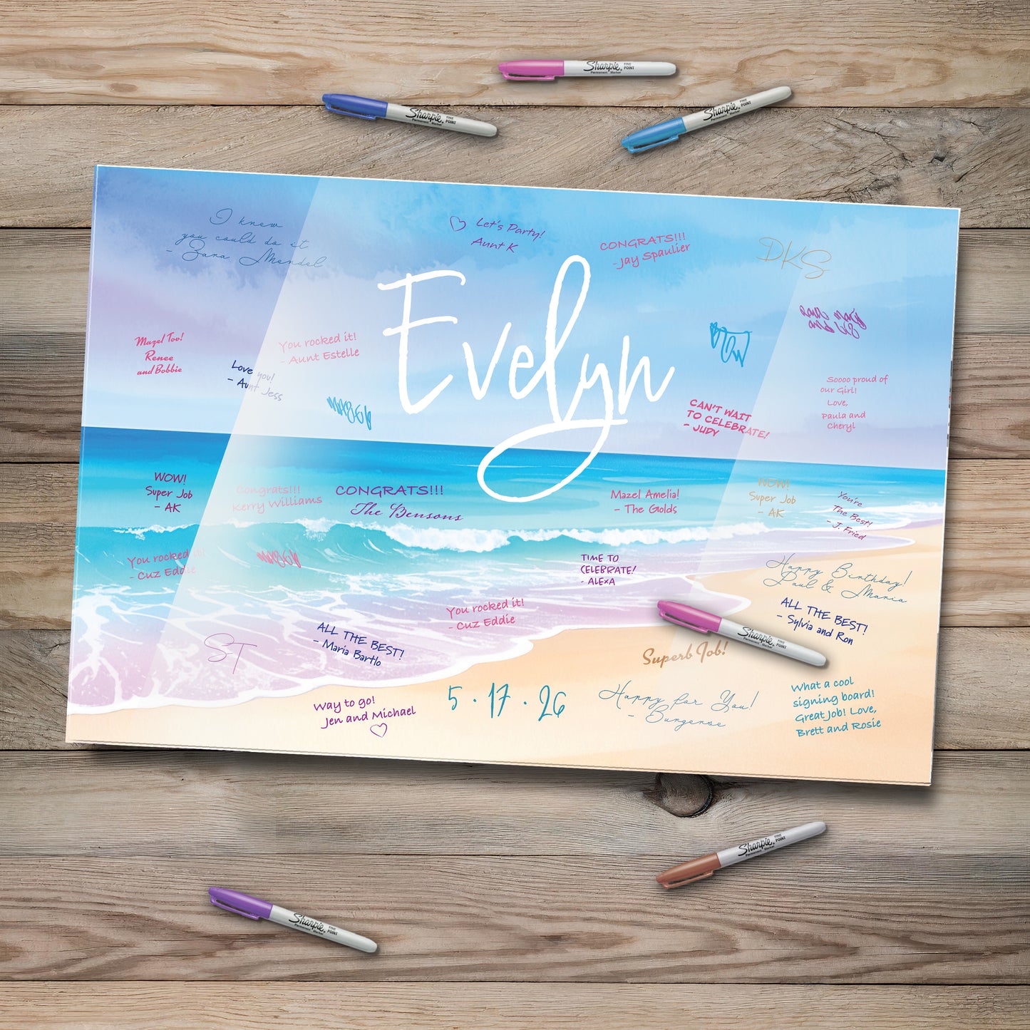 This image features a personalized beach-themed acrylic guestbook sign-in board with the name "Evelyn" elegantly written across a serene ocean backdrop. Waves gently meet the sandy shore, while colorful messages from guests surround the name. Several Sharpie markers in various colors lie nearby on a rustic wooden table, inviting attendees to leave their warm wishes.