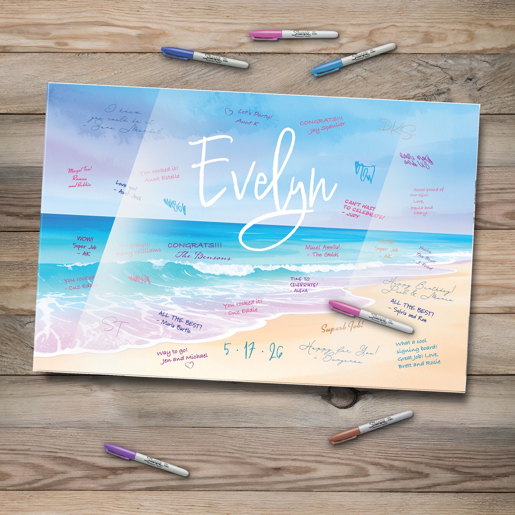 This image features a personalized beach-themed acrylic guestbook sign-in board with the name "Evelyn" elegantly written across a serene ocean backdrop. Waves gently meet the sandy shore, while colorful messages from guests surround the name. Several Sharpie markers in various colors lie nearby on a rustic wooden table, inviting attendees to leave their warm wishes.