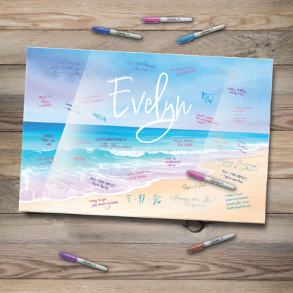This image features a personalized beach-themed acrylic guestbook sign-in board with the name "Evelyn" elegantly written across a serene ocean backdrop. Waves gently meet the sandy shore, while colorful messages from guests surround the name. Several Sharpie markers in various colors lie nearby on a rustic wooden table, inviting attendees to leave their warm wishes.