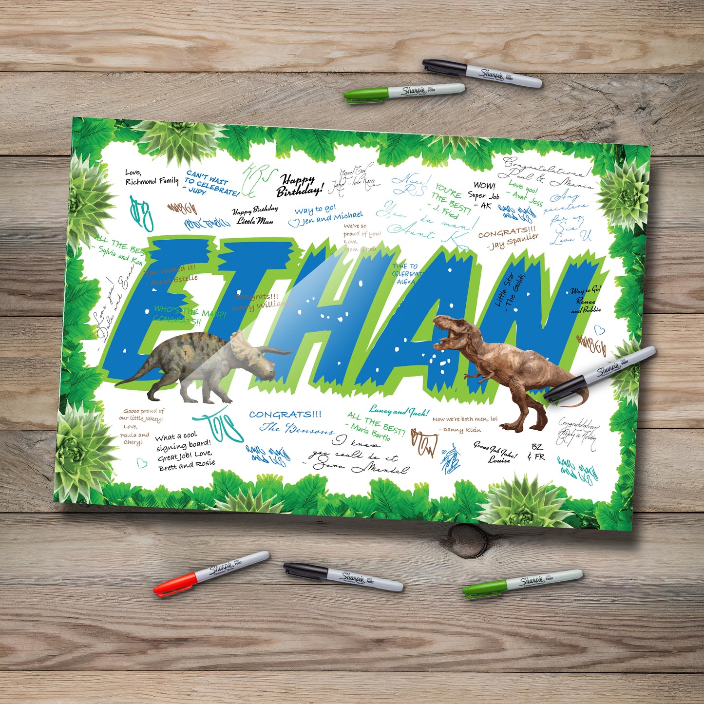 This image shows a dinosaur-themed signable acrylic board for a birthday event. The name “Ethan” is prominently displayed in large, bold blue and green letters with playful, realistic dinosaur illustrations—a triceratops and a T-rex. The border is decorated with green leaves, and the surface is filled with handwritten messages in various ink colors. The board is set on a rustic wooden table with colorful markers around it, creating a warm, festive feel