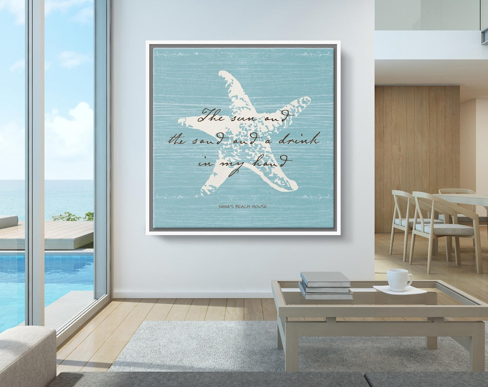 This image features a beautifully styled beach house interior with a large coastal-themed canvas on the wall. The artwork showcases a white starfish on a distressed light blue wooden background with elegant script that reads: “The sun and the sand and a drink in my hand.” Below, it’s personalized with “Nana’s Beach House.” The setting includes an ocean view, natural wood accents, and soft, neutral furnishings, creating a serene and inviting seaside atmosphere.