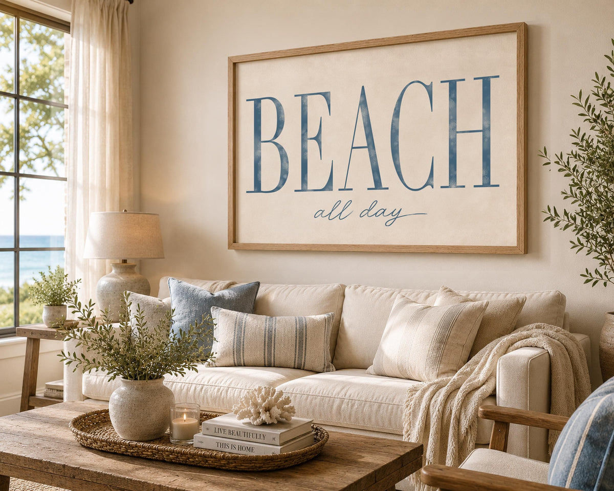 May include: A large rectangular wall art piece in a light wood frame featuring the text BEACH in large blue serif letters and the words all day in a smaller blue script font on a cream background, displayed above a beige sofa in a living room.